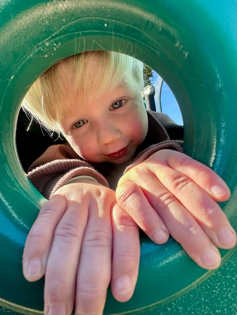 A learner peeking through a circular opening in the playground equipment, both hands resting on the lower edge