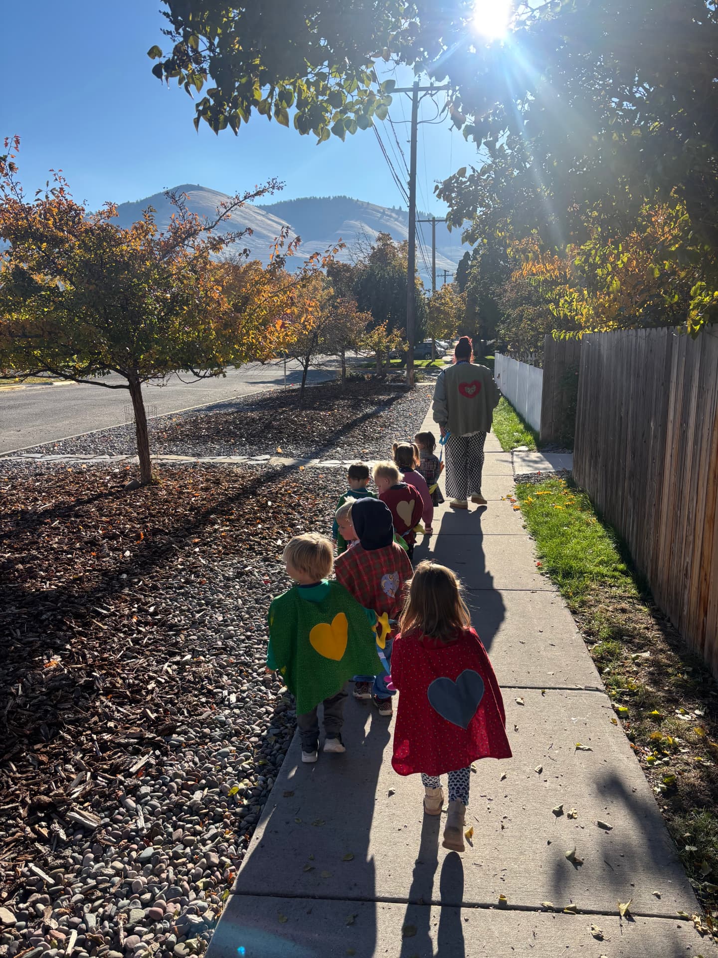 Learners in handmade heart capes walk down a sunny sidewalk with Mount Sentinel in the distance