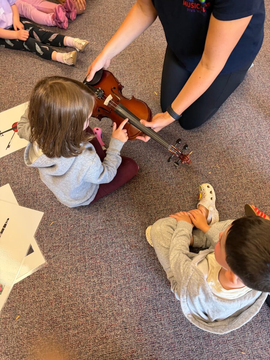 A music teacher kneels on the floor holding a small violin so a learner can explore the strings
