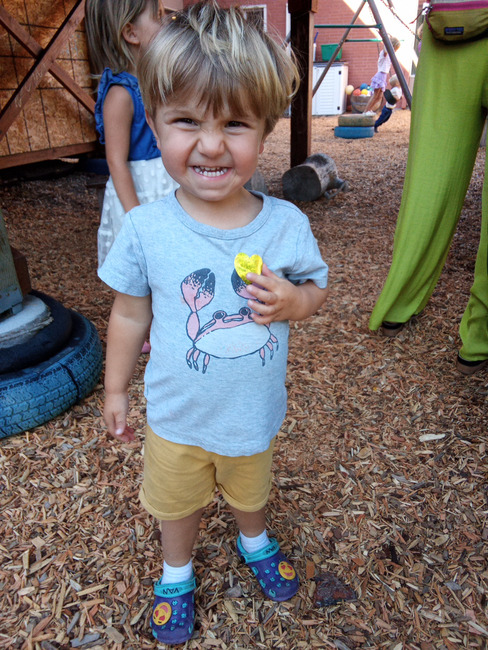 A young learner grinning on the wood-chip playground, holding up a yellow find