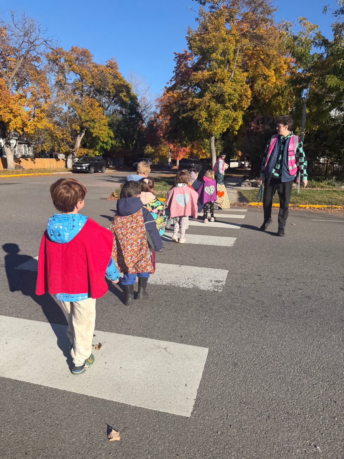 A line of caped learners holds the walking rope on a leaf-strewn sidewalk