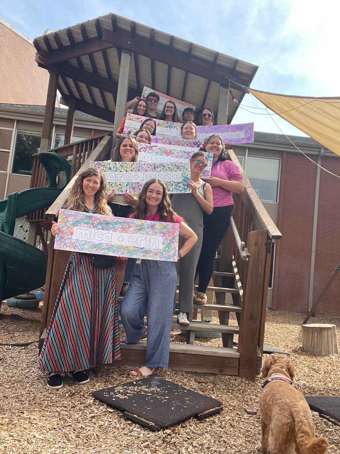 The MCS teaching team gathered on the playground steps holding hand-made name signs, with Maggie the school dog in the foreground